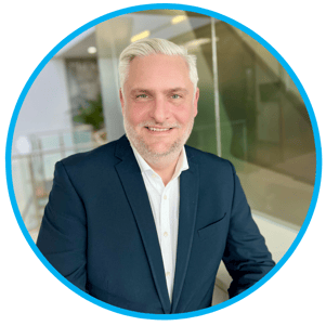 Headshot of Scott Symington CCO AJW Group, smiling wearing a navy blue blazer and white shirt, posing in a modern office setting